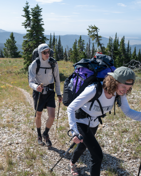 Two backpackers hike up a trail towards the camera. The front backpacker is smiling. Their packs are correctly fit.