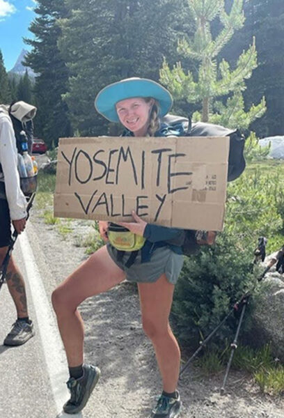 A female backpacker holds a sign that says "Yosemite Valley" with her leg extended as she attempts to hitch hike into town.