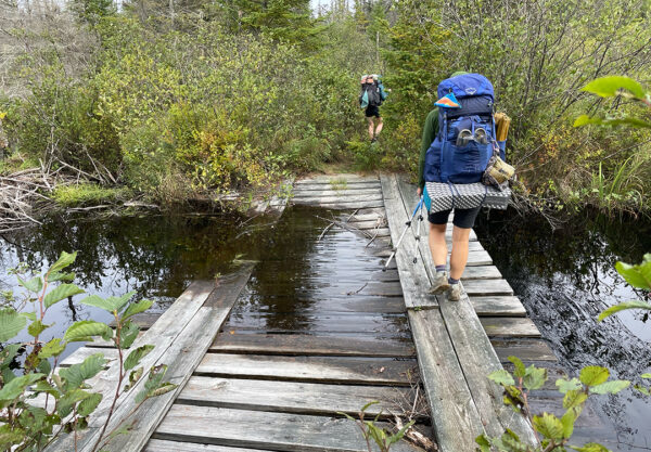 A pair of hikers cross a broken down bridge while hiking.