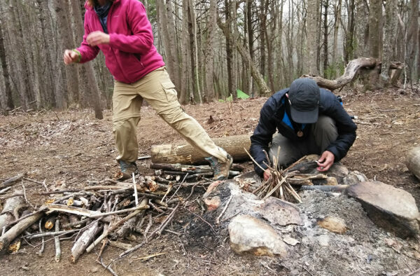 A man bends down over a fire ring while a woman in a read sweater walks away.