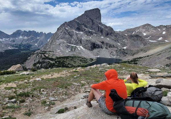 A couple of backpackers sit on a rock admiring an alpine vista