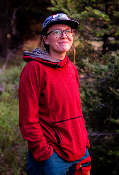 Team member Amelia smiles at the camera. She is wearing a red swaeter, glasses, and a ball cap. Amelia is a part of our production and design team.