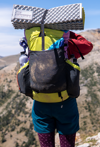 Amelia's custom ULA pack in yellow, and purple. She is hiking and her foam pad is strapped to the top of her pack.