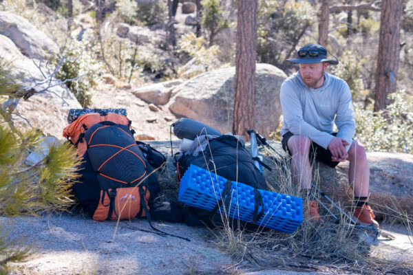 A backpacker sits on a rock, looking at two backpacks.