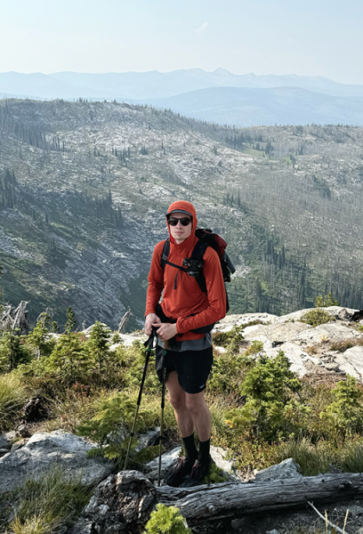 Owen testing packs on the trail. He is wearing a red hiking shirt and sunglasses. There is a mountain view behind him.