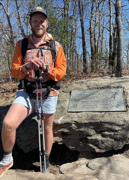 Through hiker Prof. Carl stands next to a large Appalachian Trail plaque on a rock. He is smiling.