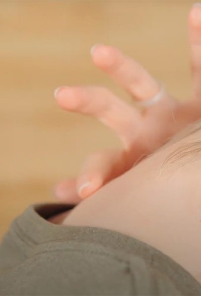 A woman points to the approximate location of the C7 vertebrae in the neck. Her head is tipped forward, and she is pointing at the lumpy bone at the base of her neck.
