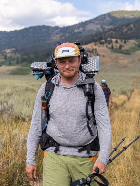 Two backpackers hike towards the camera in the Montana wilderness. The first packer is smiling; the second is blury in the background.
