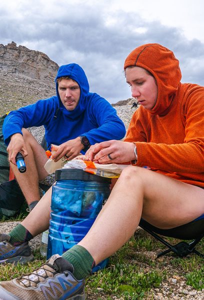 Two hikers sit down to lunch; they are using the bear canister as a table.