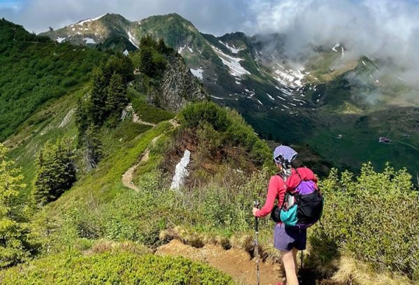 A through hiker walks along a trail with snow covered slopes behind her