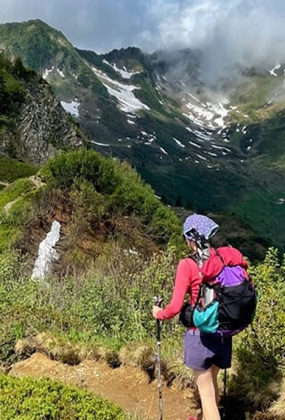 A through hiker walks along a trail with snow covered slopes behind her