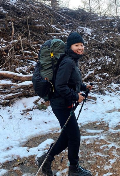 A through hiker walks through snow carrying a green ULA pack. She is wearing a hat.