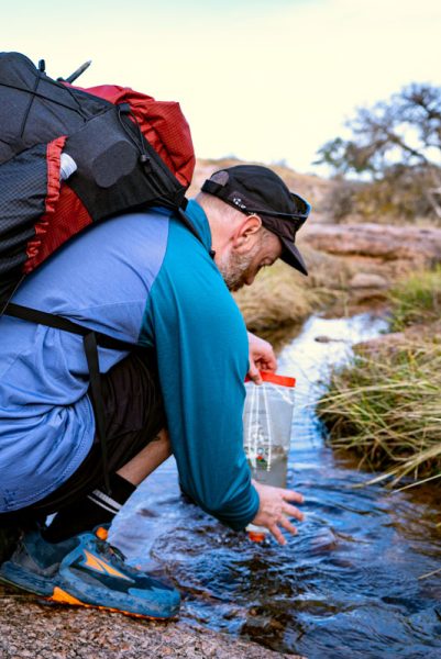 A backpacker filters water from a stream. He is wearing a ULA backpack