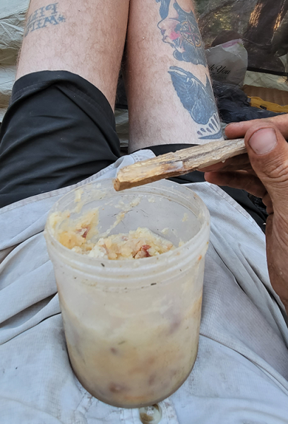 A dirty through hiker is lying down eating food out of a very dirty plastic cup and using a piece of wooden stick for a spoon.