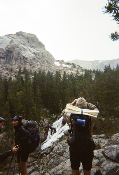 A backpacker is taking a photo of a waterfall in the rain. They are wearing rain gear. The photographer titled the image "can't share rain gear." Very true.