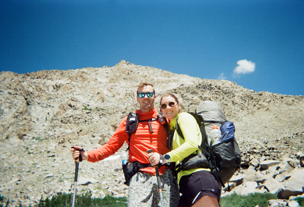 A couple smiles at the camera with a mountain behind them.