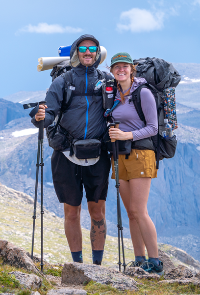 A photo of Garrett and Rebekah standing on a mountain top and smiling for the camera. They are holding trekking poles and wearing ULA packs.