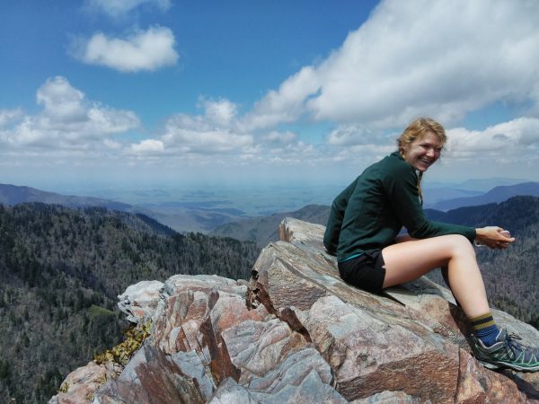 A smiling female hiker sits on a rock cropping at the top of a mountain. Behind her is a large vista of rolling hills. The sky is blue with puffy clouds.