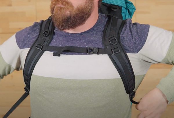 Image shows a male in a pack with S-strap shoulder straps. He is tightening the straps.