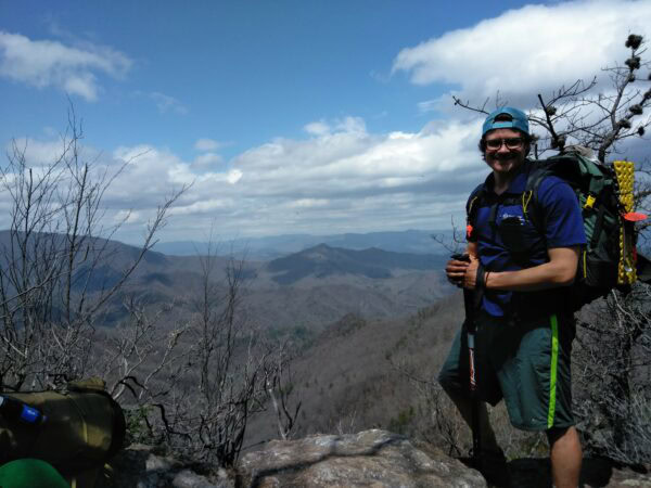 A backpacker, wearing a green robic ULA pack, stands on the top of a mountain.