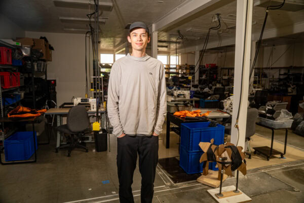 A portrait of Owen, a vital member of the ULA team in Logan, Utah. He is wearing a grey hat and shirt, and black pants. His hands are in his pockets.
