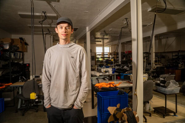 A portrait of Owen, a vital member of the ULA team in Logan, Utah. He is wearing a grey hat and shirt, and black pants. His hands are in his pockets.